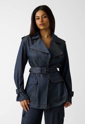 Frau trägt dunkelblaue Jeansjacke mit Gürtel und großen Revers sowie passende Hose mit Taschen, steht vor neutralem Hintergrund.