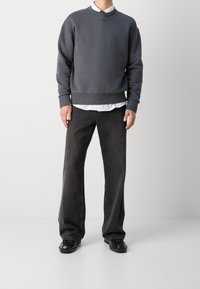 Felpa grigia con colletto a girocollo, polsini e orlo a costine; indossata sopra una camicia bianca. Jeans grigio scuro a gamba larga e scarpe nere completano l'outfit.
