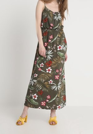 Vrouw die een mouwloze olijfgroene maxi-jurk met tropische bloemenprint en gele sandalen met hakken draagt, staand tegen een effen achtergrond.