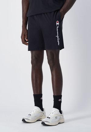 Shorts sportivi neri con una leggera lucentezza, con il logo "Champion" in bianco sul lato sinistro. Abbinati a sneakers bianche e calze nere.