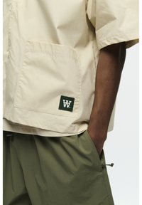 Beige Kurzarmjacke mit einer Brusttasche und grünem Logopatch, kombiniert mit lockeren olivgrünen Shorts mit seitlichem Kordelzug.
