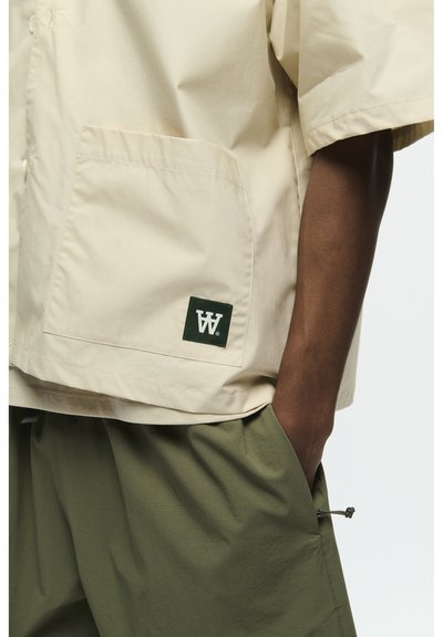 Veste beige à manches courtes avec une poche poitrine et un écusson logo vert, associée à un short ample vert olive avec un cordon de serrage sur le côté.