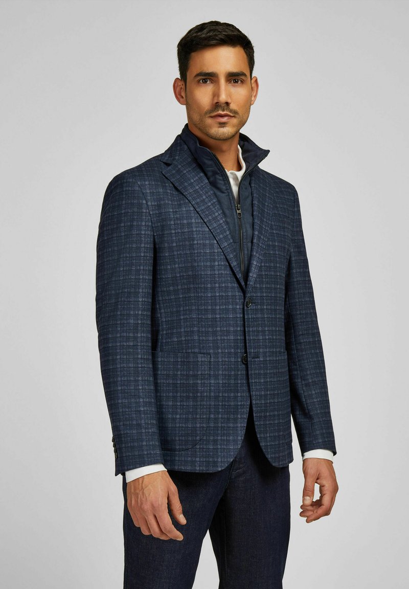 Marineblau kariertes Blazer mit strukturierten Schultern, Revers und zwei Taschen. Über einer dunklen Zip-Jacke mit Stehkragen getragen.