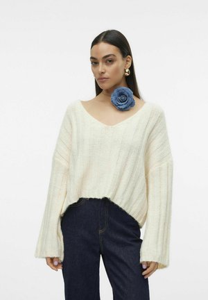 Krämfärgad, ribbad stickad tröja med djup V-ringning och vida ärmar. Har en blå tygros nålad på kragen. Mörka jeans.
