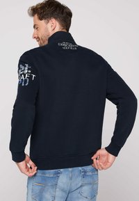 Sudadera azul marino con cuello alto, puños acanalados y gráficos detallados en la manga y la parte posterior. Fabricada con un tejido suave y texturizado.