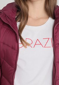Burgundy puffy jacket with a zippered front and quilted texture, layered over a white t-shirt featuring red text "GRAZI."