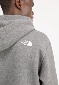 Szara bluza z kapturem, wykonana z miękkiego materiału, z białym logo The North Face na plecach. Projekt zawiera przestronny kaptur i ściągacze.