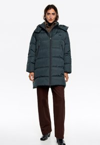 Ciemnozielona puchowa kurtka o pudełkowym kroju, z zamkiem błyskawicznym z przodu, kapturem i bocznymi kieszeniami. Tkanina o fakturze z poziomymi szwami. Połączona z brązowymi spodniami.