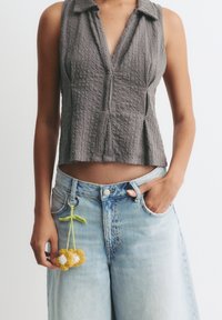 Grauer ärmelloser Oberteil aus strukturiertem, geripptem Stoff mit Kragen; kombiniert mit hellblauen hochgeschnittenen Jeans und einem farbenfrohen, floralen Schlüsselanhänger.