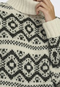 Pull crème avec un col montant côtelé, présentant des motifs géométriques noirs complexes et une finition en maille texturée sur l'ensemble.