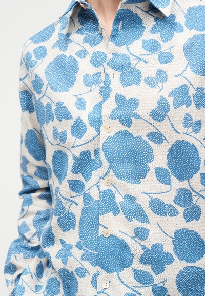 Λευκό πουκάμισο με κουμπιά, με μπλε ανθισμένο σχέδιο που περιλαμβάνει μεγάλα λουλούδια και φύλλα, φορεμένο από άτομο με το ένα χέρι στην τσέπη.
