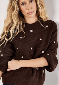 Maglione lavorato a maglia marrone con una vestibilità rilassata, caratterizzato da piccole ricami floreali bianchi sparsi sul tessuto e maniche arrotolate.