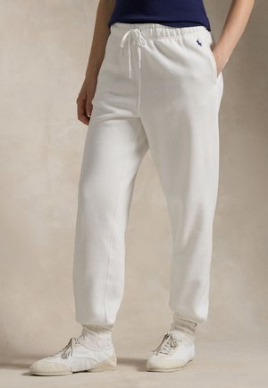 Personne portant un pantalon de survêtement blanc à cordon, des chaussettes blanches et des chaussures décontractées blanches, debout avec une main dans la poche.