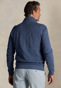 Marineblauwe gebreide trui met een hoge kraag, geribbelde manchetten en een gladde textuur. Gedragen over lichtgekleurde broeken met knoopaccenten.