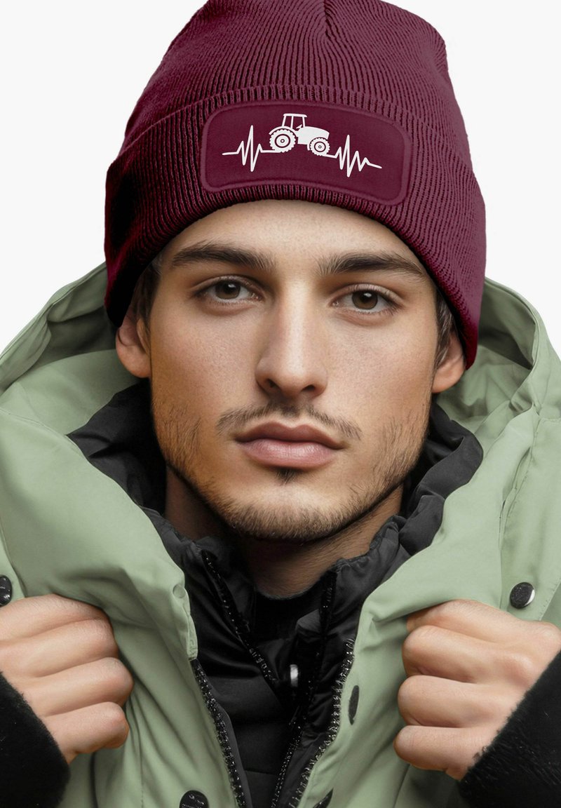 Burgundy knit beanie with tractor graphic and heartbeat line patch; paired with a green, puffy jacket and black inner layer.