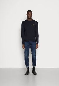 Maglione a maglia blu navy con collo rotondo e logo giallo, abbinato a jeans blu e stivali neri alla caviglia; in piedi davanti a uno sfondo uniforme.