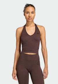 Hnědý sportovní crop top s výstřihem do V a tenkými ramínky, v kombinaci s vysokými legínami se stejným vzorem. Jemný detail loga je viditelný.