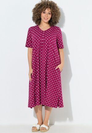 Kobieta z kręconymi włosami, ubrane w różową sukienkę do kolan z białymi gwiazdkami i białe sandały wsuwane, stoi przy białej ścianie.