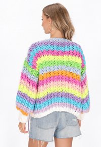 Daudzkrāsains adīts džemperis ar horizontālām svītrām pasteļtoņos, ar brīviem piedurknēm un masīvu tekstūru; valkāts virs džinsu šortiem.