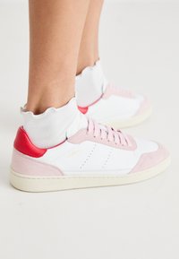 Hvite sneakers med rosa semsket tå og hæl detaljer, rød hælflik, og en teksturert hvit ankelstrømpe med bølget kant.