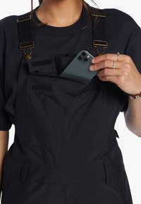 Osoba oblečená v černých overalách a černém tričku si vkládá tmavý smartphone do přední kapsy na hrudi overalu.