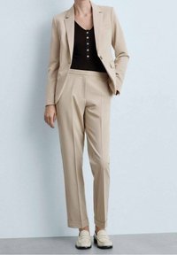 Costume beige sur mesure avec une veste cintrée et un pantalon droit. La tenue comprend un haut noir et des mocassins crème. Tissu lisse avec des lignes épurées.