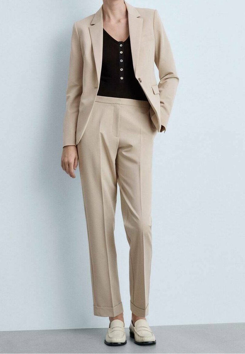 Costume beige sur mesure avec une veste cintrée et un pantalon droit. La tenue comprend un haut noir et des mocassins crème. Tissu lisse avec des lignes épurées.