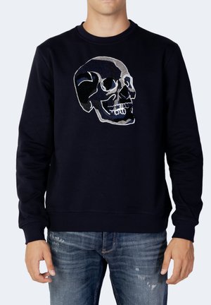 Mand iført sort sweatshirt med stort broderet kraniedesign, kombineret med blå slidte jeans mod ensfarvet baggrund.
