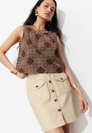 Femme portant un haut marron sans manches à motif, jupe beige à boutons sur le devant avec poches, bracelets dorés et bijoux assortis, posant contre un mur blanc.