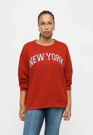 Ung person med flettet hår og briller iført en rød sweatshirt med teksten "NEW YORK CITY" og blå jeans, stående foran en ensfarvet baggrund.