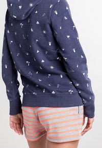 Felpa navy con motivi bianchi di icone tropicali e forme geometriche; abbinata a pantaloni corti grigi con strisce arancioni.