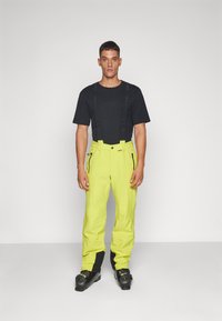 Gele waterdichte skibroek met verstelbare bretels, ritsen op de zakken, gedragen met een zwart t-shirt en zwarte skischoenen.