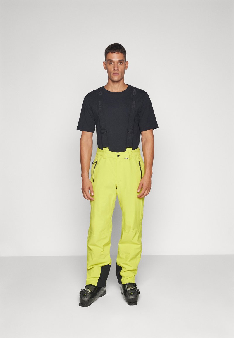 Gele waterdichte skibroek met verstelbare bretels, ritsen op de zakken, gedragen met een zwart t-shirt en zwarte skischoenen.