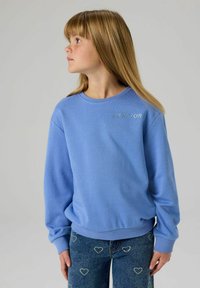 Felpa azzurro chiaro con polsini e colletto a coste; presenta la scritta 'NEW YORK' in testo argentato scintillante. Abbinata a jeans blu con motivo a cuori.