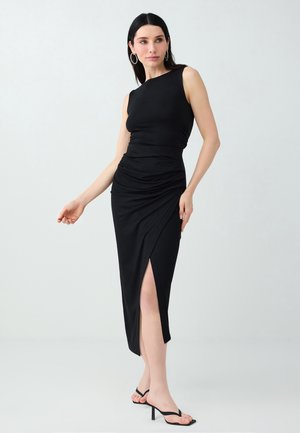 Femme aux cheveux noirs portant une robe noire sans manches fendue sur le côté et des sandales noires à talons, debout contre un fond clair uni.