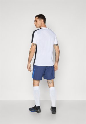 Sportstøj bestående af en hvid og sort kortærmet top samt blå shorts med en kontrasterende blå detalje, parret med sorte stødder og hvide sokker.