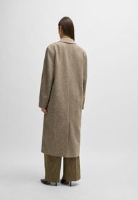 Long, brown coat with a textured finish and light speckles, featuring a notch collar and dropped shoulders, paired with wide plaid trousers.