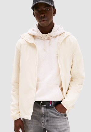 Giacca zip-up crema chiaro con cappuccio, indossata sopra un hoodie beige chiaro. Jeans grigi e una cintura nera con accenti rossi, bianchi e blu.