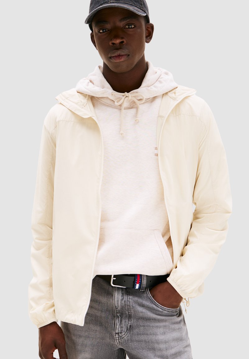 Giacca zip-up crema chiaro con cappuccio, indossata sopra un hoodie beige chiaro. Jeans grigi e una cintura nera con accenti rossi, bianchi e blu.