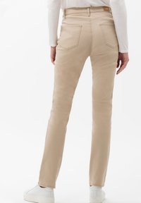 Pantalones beige ajustados hechos de tela suave, con una cintura de altura media y bolsillos traseros. Combinados con zapatillas blancas frente a un fondo sencillo.