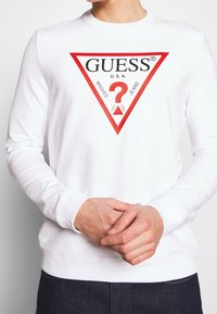 Sudadera blanca de manga larga con un gran logo triangular rojo que lleva "GUESS® U.S.A" en negro y un signo de interrogación en el centro.