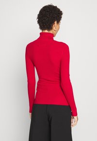 Femme aux cheveux courts et bouclés portant un pull à col roulé rouge côtelé ajusté et un pantalon large noir, vue de dos.