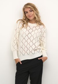 Weißer Strickpullover mit Rautenmuster, geripptem Rundhalskragen und Bündchen. Kombiniert mit schwarzen Hosen in lockerer Passform.
