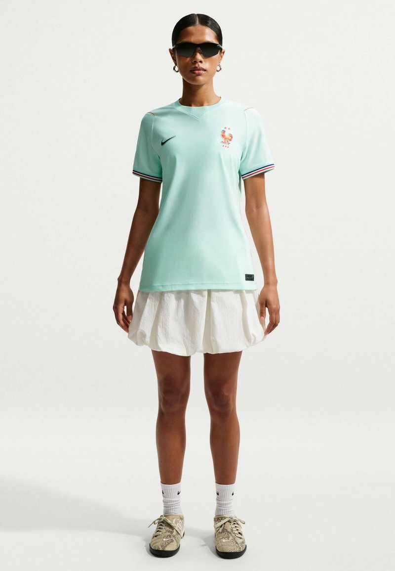 Donna in piedi che indossa la maglia della nazionale di calcio francese azzurro chiaro, gonna bianca, calzini bianchi Nike, scarpe beige e occhiali da sole neri su sfondo semplice.