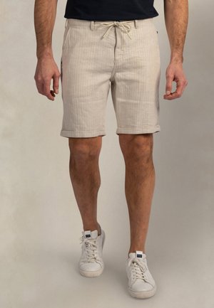 Man draagt beige knielange korte broek met een trekkoord in de taille en witte sneakers, staand tegen een effen achtergrond.