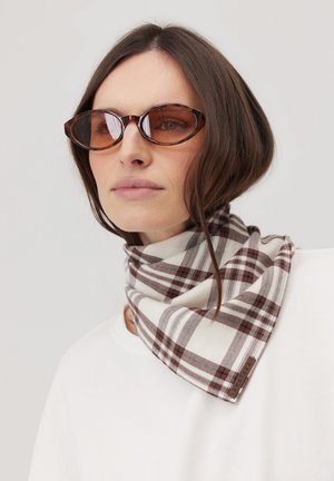 Kobieta z krótkimi brązowymi włosami, nosząca owalne okulary przeciwsłoneczne z bursztynowym odcieniem i biało-brązowy kraciasty szalik na białym topie na jednolitym tle.