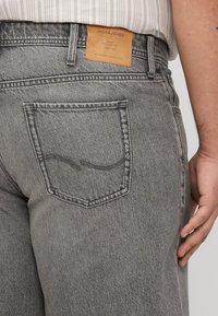 Jeans din denim gri cu o finisare texturată, având un buzunar la spate cu o cusătură curbată și o etichetă din piele maro deschis inscripționată cu "JACK & JONES."