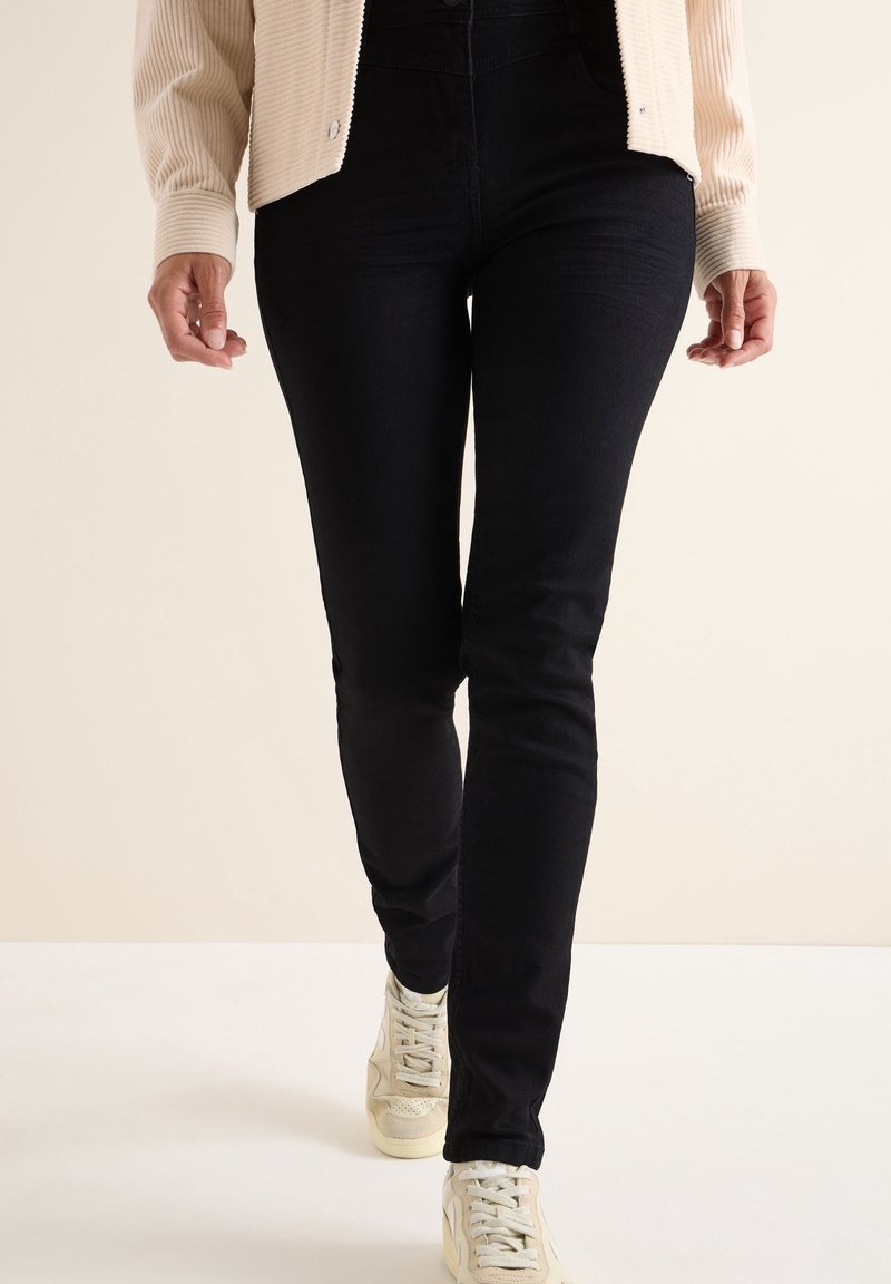Personne portant un jean skinny noir, une veste en velours côtelé crème et des baskets blanches marchant vers l'avant sur un fond uni.