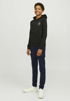 Sweatshirt noir à capuche avec poche frontale et logo, associé à un pantalon cargo bleu marine avec poches latérales, et des baskets blanches.