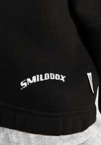 Tissu noir avec texte brodé blanc "SMILODOX" et une petite étiquette en tissu blanc sur le côté droit du vêtement.